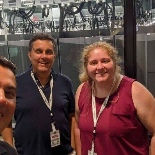 Three people wearing ID badges stand in front of a window, behind which are visible server racks and cables in a high-tech data center environment.